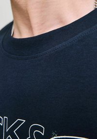 Tamsiai mėlynos medvilnės marškinėliai su guma apykakle, su baltu ir oranžiniu grafikiniu atspaudu, turinčiu tekstūruotą, lygų paviršių.