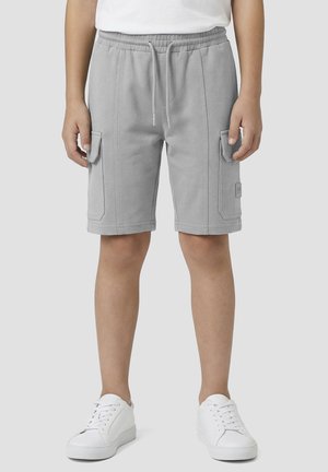 Bambino che indossa pantaloncini cargo grigio chiaro con coulisse in vita e tasche laterali, abbinati a scarpe da ginnastica bianche e una camicia bianca.