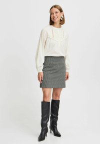 Weiße Bluse mit strukturierten Details, langen Ärmeln und einer Knopfleiste, kombiniert mit einem grauen karierter Minirock und knielangen schwarzen Stiefeln.