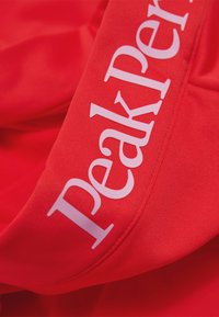 Rode stof met een gladde, elastische textuur en een prominent wit "Peak Performance" logo aan de zijkant. Ontworpen voor actief gebruik.