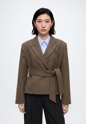 Jeune femme aux cheveux courts noirs portant un blazer marron ceinturé sur une chemise bleu clair et un pantalon noir, faisant face à l’avant sur un fond uni.