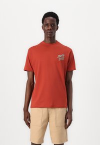 Red cotton T-shirt with short sleeves and a crew neck, featuring a small "Santa Cruz" logo in grey on the left chest. Worn with beige shorts.