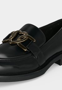 Μαύρο δερμάτινο loafer με χοντρή σόλα και μεγάλο χρυσό μεταλλικό αλληλοσυνδεόμενο αλυσίδα στην κορυφή.