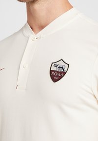 Kremowa koszulka polo z dwoma guzikami na dół, z haftowanym herbem AS Roma w bordowym kolorze po lewej stronie klatki piersiowej. Teksturowana tkanina.