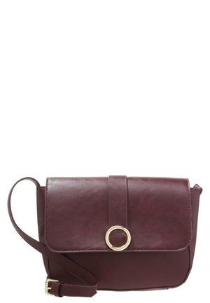 Sac bandoulière en cuir marron foncé avec un rabat frontal fixé par une sangle et un ornement circulaire doré, ainsi qu'une bandoulière réglable.