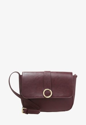 Sac bandoulière en cuir marron foncé avec un rabat frontal fixé par une sangle et un ornement circulaire doré, ainsi qu'une bandoulière réglable.