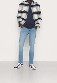 Kostkovaná flanelová košile v černé, bílé a modré barvě, přeložená přes tmavě modré tričko. Světle modré džíny a tmavě modré tenisky s bílými akcenty dokončují vzhled.