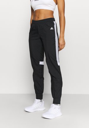 Athletic person wearing black Adidas track pants with gray stripes, white Adidas crop top, white Adidas sneakers, standing on gray floor.