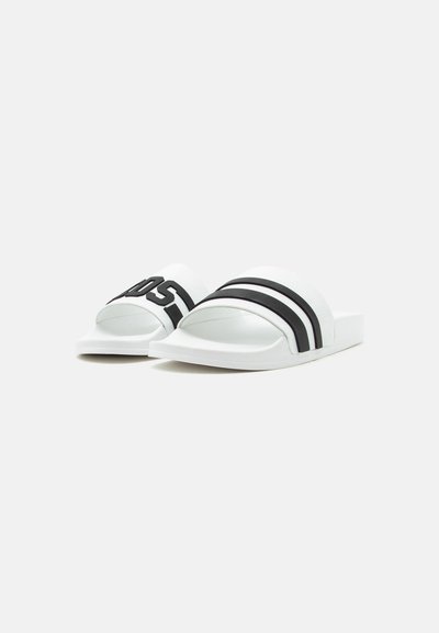 Sandales blanches à semelle plate avec deux bandes noires sur l'une et des lettres noires en gras sur l'autre, présentées sur un fond clair uni.