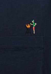  Tela de color azul marino con un bolsillo que presenta un diseño bordado de una figura asando a la parrilla con llamas naranjas sobre una parrilla de carbón negra.