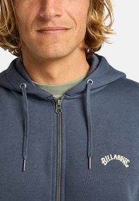 Marineblå zip-up hættetrøje med snore og broderet "BILLABONG" logo. Glat, blød tekstur; metallynlås og accentdetaljer til stede.