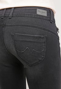 Vaqueros de denim negro con un diseño distintivo de bolsillo trasero curvado, con costuras sutiles y un parche de cuero con la marca en la cintura.