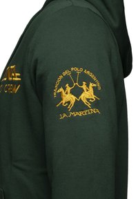 Sudadera verde con bordado amarillo de jugadores de polo, el texto "TRADICIÓN DEL POLO ARGENTINO" y "LA MARTINA" arriba. Textura de tejido suave.