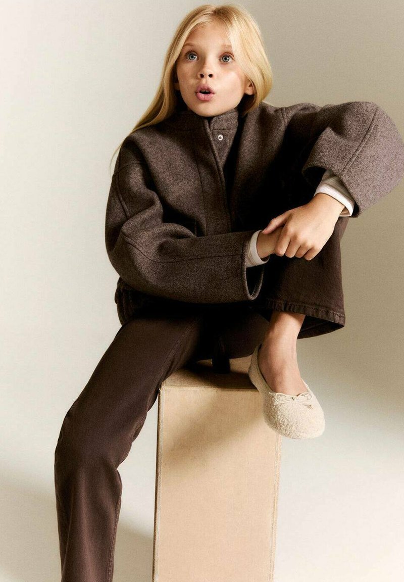 Bruine wollen oversized jas, beige coltrui, donkerbruine broek, lichtgekleurde instappers met strikaccenten, zittend op een doos.