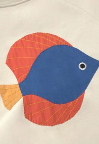 Poisson en tissu bleu aux nageoires et queue orange, détaillé avec des coutures jaunes, cousu sur un fond en tissu crème.