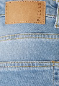 Tecido de denim azul claro com costura amarela, apresentando uma etiqueta de couro castanho na cintura com "PIECES" gravado.