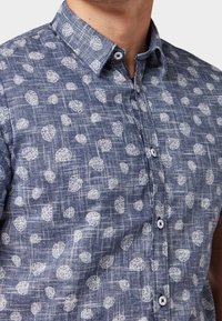 Camicetta a maniche corte con bottoni in chambray blu con un motivo a foglie bianche. Presenta colletto e patta dei bottoni frontale.