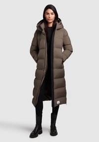 Lång brun puffjacka med huva, utrustad med dragkedja och sidofickor. Texturerad tyg med ett quiltat mönster och svarta detaljer.