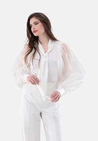 Blusa bianca con maniche a sbuffo trasparenti, colletto con lacci e polsini con bottoni, abbinata a pantaloni bianchi a vita alta. Tessuto liscio e morbido.