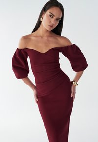 Abito borgogna off-the-shoulder con tessuto testurizzato, corpino aderente e maniche a sbuffo fluide. Un bracciale dorato accentua il polso.