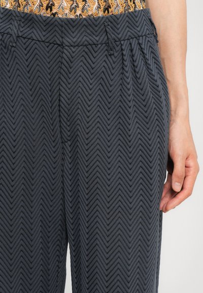 Pantaloni de culoare gri închis, cu un model în zig-zag, confecționați dintr-un material texturat, având un elastic cu pliseuri subtile și o mână odihnindu-se aproape de lateral.