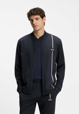 Junger Mann trägt schwarze Jacke mit Reißverschluss und passende Hose mit weißen vertikalen Streifen und "BOSS"-Logos, Hände in den Taschen.