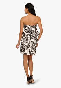 Femme vêtue d'une robe blanche sans bretelles avec un grand imprimé floral noir et un détail volanté à l'arrière, associée à des chaussures à talons hauts noires.