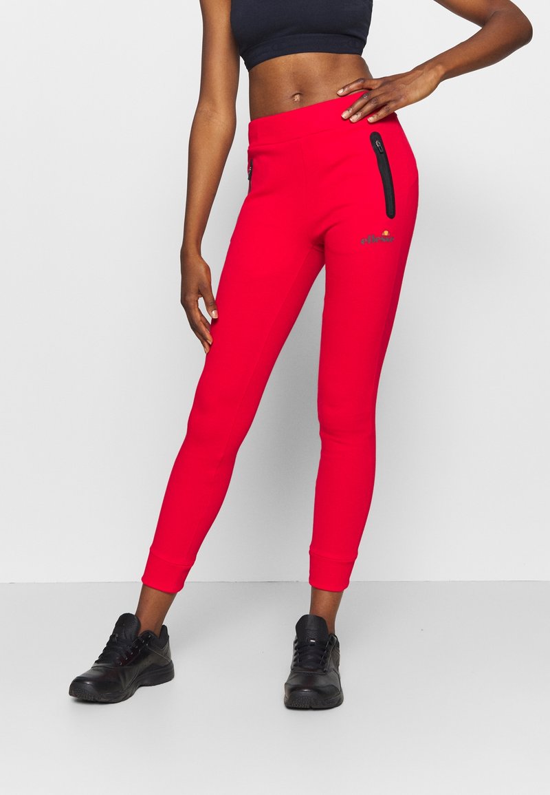 Femme portant un pantalon de jogging ajusté rouge vif avec des poches zippées noires, des baskets noires et un crop top noir sur un fond blanc uni.