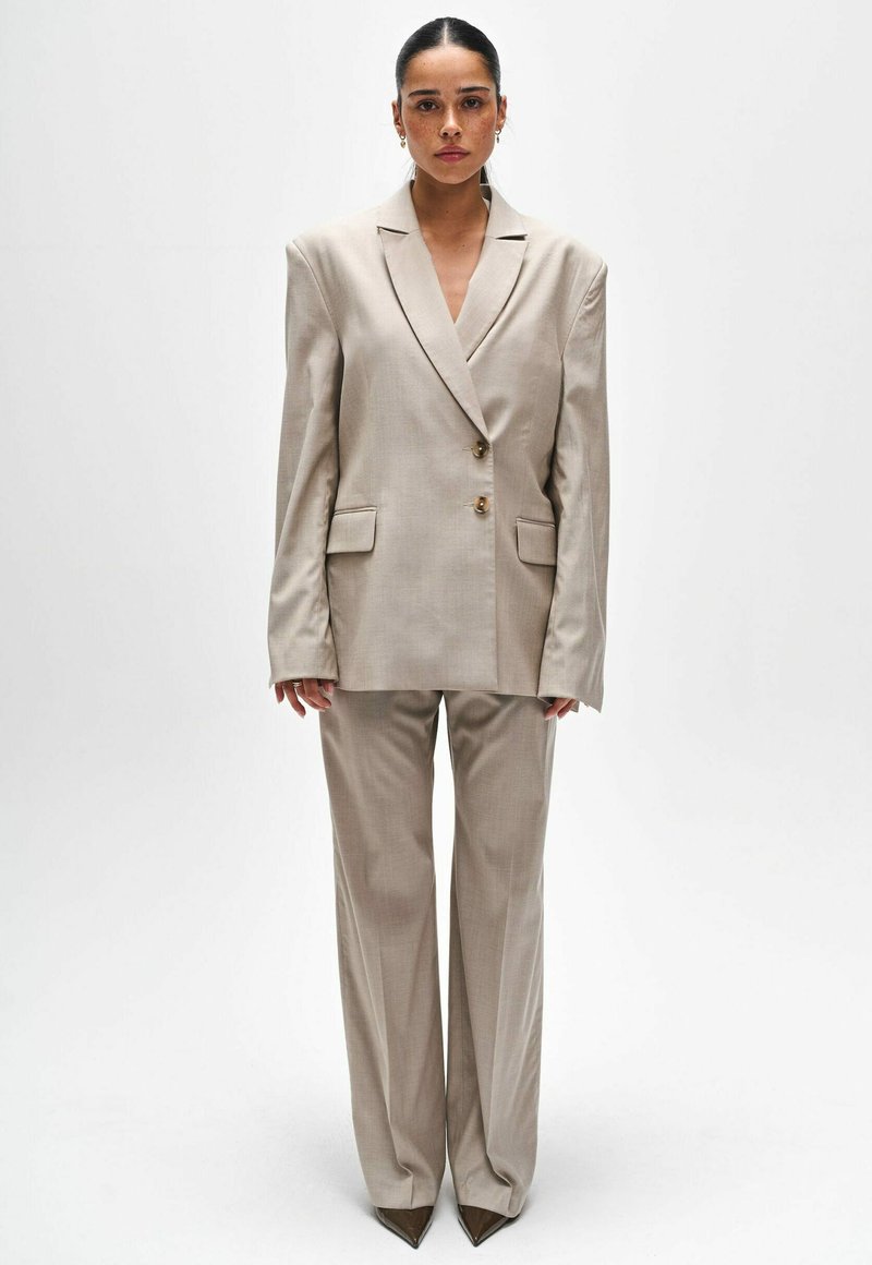 Beige op maat gemaakt pak met een oversized blazer met twee knopen en gelobde revers, gecombineerd met losse broek en puntige schoenen.