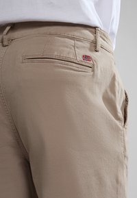 Pantaloni beige con una texture liscia, dotati di una tasca posteriore e di una piccola bandiera norvegese ricamata sopra la tasca.