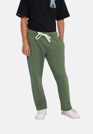 Mann trägt olivgrüne Hose mit Kordelzug und Seitentaschen, kombiniert mit weißen Sneakers und ein schwarzes kurzärmeliges Hemd.