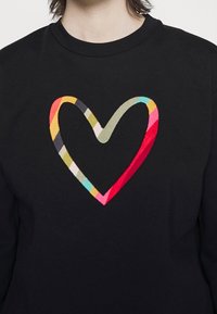 Personne portant un t-shirt noir avec un grand cœur rayé multicolore centré sur la poitrine.