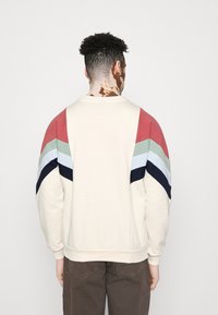 Felpa con collo rotondo di colore crema con motivo a zigzag multicolore in rosso, verde, blu e blu navy sulle maniche. Tessuto morbido, vestibilità rilassata.