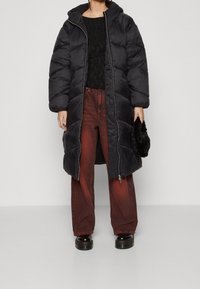 Svart puffjacka med huva, dragkedja framtill och pösig kviltad textur; kombinerad med mörkröda denimbyxor och svarta platåskor.