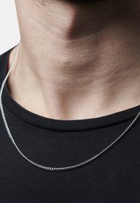 Silverkedja som vilar på en bar nacke, med en vriden design och en slät yta, med en svart t-shirt med rund halsringning i bakgrunden.