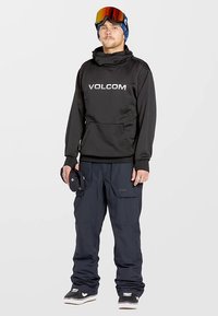 Black Volcom hoodie with silver logo, paired with navy snow pants featuring pockets. The model wears a blue beanie and reflective ski goggles.