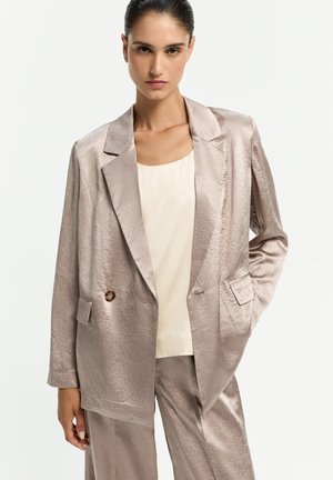 Frau trägt einen locker sitzenden, glänzenden beigen Anzug mit einreihiger Jacke und passenden weit geschnittenen Hosen, kombiniert mit einem cremefarbenen Oberteil.