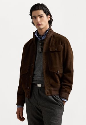 Braune Wildlederjacke mit frontalen Taschen, geripptem Kragen und Bündchen. Genossen wird sie über einem grauen Pullover und einem hellblauen Hemd mit gepunktetem Muster.