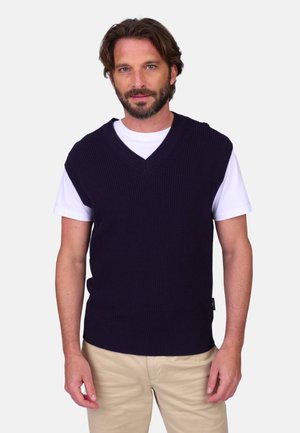 Homme portant un pull sans manches en maille bleu marine par-dessus une chemise blanche à manches courtes et un pantalon beige, debout devant un fond uni.