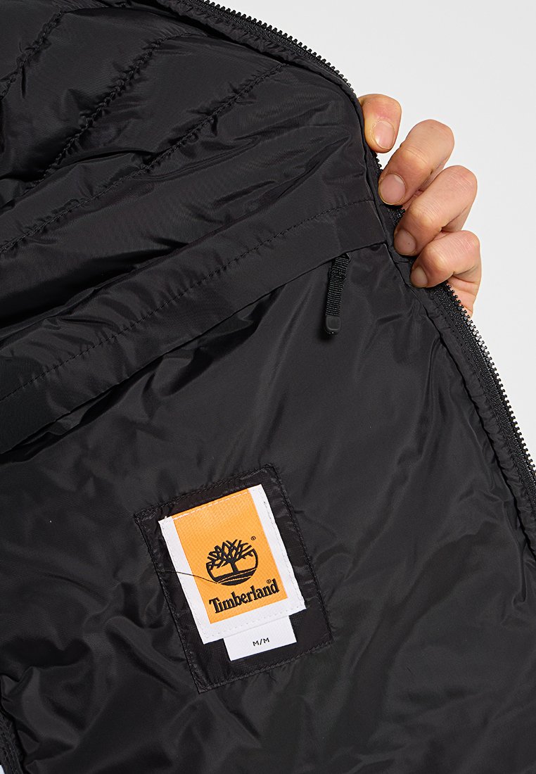 Czarna izolowana kurtka z pikowanym wnętrzem, z etykietą Timberland i kieszenią na zamek. Gładka tekstura nylonowa.