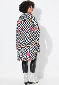 Zwart-wit geruite capuchon jurk met roze en oranje sterren en vredessymbolen, gedragen met glanzende zwarte leggings en metallic zilveren laarzen.