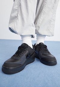 Henkilö, joka on pukeutunut mustiin Nike Air Force 1 -tennareihin, valkoisiin sukkiin ja vaaleanharmaisiin leveisiin housuihin, seisoo vaaleansinisen lattian päällä.