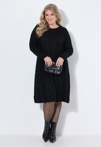 Abito nero lavorato a maglia con maniche lunghe e pattern a trecce, abbinato a una borsa a tracolla nera con texture e stivali alla caviglia con borchie.