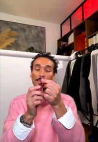 Persona con un maglione rosa applica rossetto rosso in una stanza con un appendiabiti, una libreria e un'opera d'arte astratta sul muro sullo sfondo.