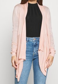 Ljusrosa cardigan med öppen framtid, ribbade ärmar och mjuk textur. Kombineras med en figursydd svart topp och blå jeans.