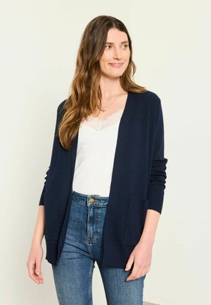 Woman with long brown hair wearing a navy blue cardigan over a white lace-trimmed top and blue jeans, looking to the side and smiling slightly.
