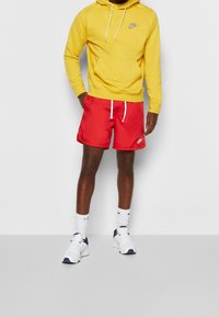 Muž v žluté mikině Nike, červených šortkách Nike s bílými šňůrkami, bílých ponožkách Nike a bílých teniskách s námořnickými akcenty.