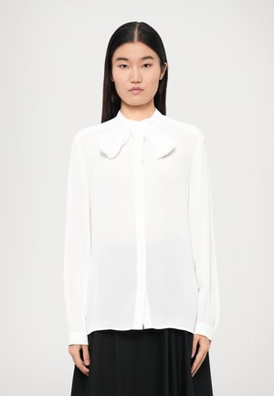 Blouse blanche à manches longues avec un nœud au col, en tissu doux, coupe ajustée, boutonnage sur le devant, et poignets avec une finition soignée.