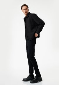 Schwarze Jacke mit Kragen, Brusttasche, über einem schwarzen gerippten Pullover getragen, kombiniert mit schwarzen Slim-Fit-Hosen und schwarzen Schuhen.