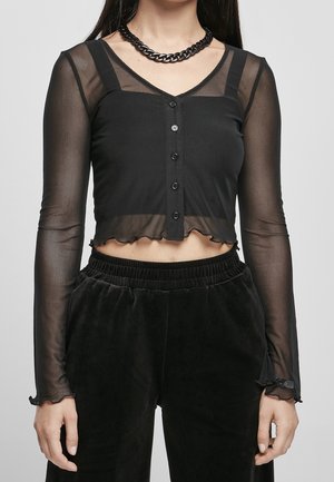 Frau trägt ein schwarzes, durchsichtiges Croptop mit langen Ärmeln, Knöpfen und gerafften Kanten, kombiniert mit schwarzen Hose mit hoher Taille und einer schwarzen Halskette mit Kette.
